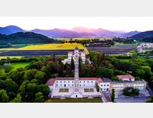 Veduta aerea di un complesso storico con villa centrale e cupola, collegato da una lunga scalinata a un edificio più basso con tetti in tegole e cortili. Intorno si estendono alberi e campi coltivati a fasce, con colline e montagne sullo sfondo.