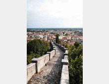 Camminamento in pietra con pavimentazione a ciottoli e muretti bassi, che scende lungo una collina tra la vegetazione. Sullo sfondo si vede un’ampia vista panoramica della città con tetti in terracotta, campanili e edifici fino all’orizzonte.