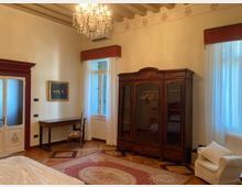 Interno di una camera arredato in stile classico, con armadio in legno scuro, scrittoio con sedia e lampadario di cristallo sotto un soffitto con travi a vista. Due grandi finestre con tende bianche e mantovane rosse illuminano un pavimento in parquet con tappeto e una poltrona chiara.