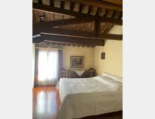 Camera da letto in stile rustico con soffitto a travi di legno a vista e pavimento in legno lucido. Un letto matrimoniale con biancheria bianca è affiancato da una finestra con tende e da una cassettiera con quadro e sedie lungo la parete.