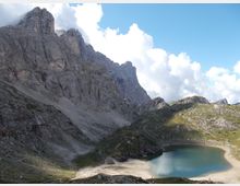 Paesaggio montano con alte pareti rocciose a strapiombo e un lago alpino turchese in una conca, circondato da ghiaioni e pendii erbosi. Sullo sfondo cielo azzurro con nuvole e crinali rocciosi.