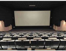 Sala cinematografica vuota con file di poltrone grigie disposte in pendenza e un grande schermo bianco frontale. Pareti e soffitto scuri con illuminazione laterale e uscite di sicurezza ai lati.