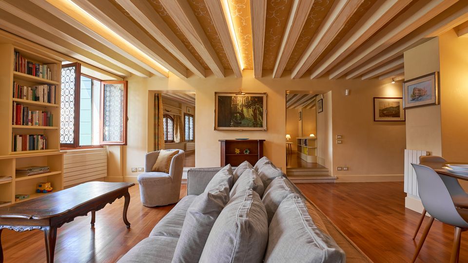 Ampio soggiorno di un appartamento con soffitto a travi in legno a vista, pavimento in parquet e illuminazione a incasso. Nella stanza si vedono una libreria a parete, finestra con inferriata decorativa, divano e poltrona, tavolino, quadri alle pareti e un’area pranzo con tavolo e sedie.