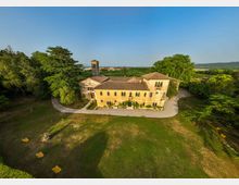 Veduta aerea di una villa storica in pietra con tetto in coppi e piccola torre campanaria, circondata da un ampio prato e da alberi ad alto fusto. Un vialetto curva attorno all’edificio e sullo sfondo si vedono campi e colline sotto un cielo sereno.
