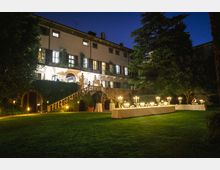 Villa storica con facciata illuminata e ampie finestre, circondata da un giardino curato. In primo piano, tavoli apparecchiati con candelabri e decorazioni, disposti sull'erba verde sotto un cielo serale.