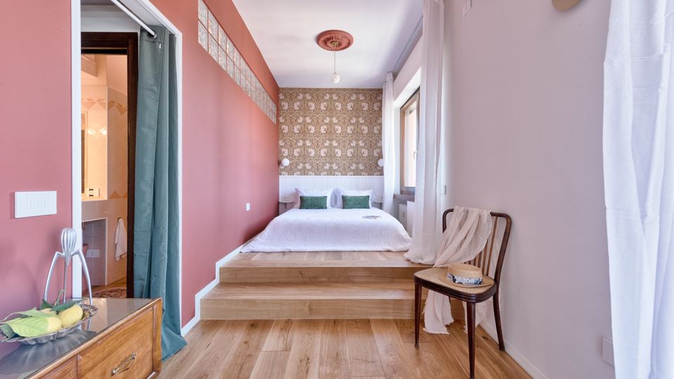 Camera da letto luminosa con letto su pedana rialzata e parete di fondo con carta da parati decorata e testiera bianca. Pavimento in legno, pareti rosa, finestra con tende bianche e accesso al bagno tramite apertura con tenda sul lato sinistro.