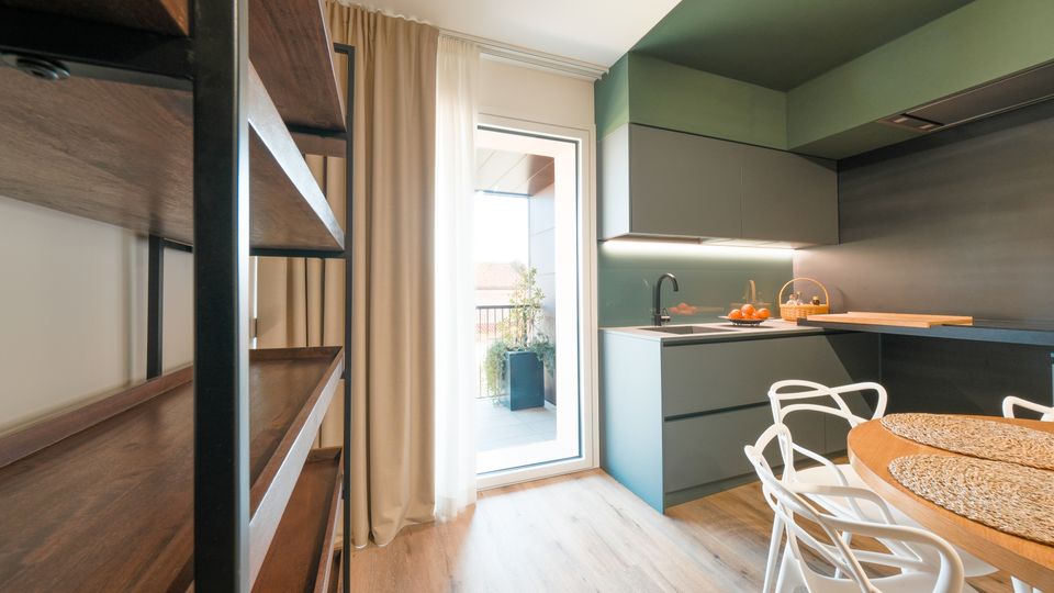 Cucina moderna con mobili verde salvia e grigi, piano di lavoro con lavello e illuminazione sottopensile, affiancata da un tavolo rotondo in legno con sedie bianche. Una portafinestra con tende chiare si apre su un balcone con vaso e ringhiera, mentre a sinistra si vede una libreria in legno e metallo.