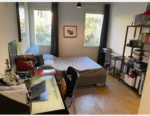 Camera da letto con scrivania e computer sulla sinistra, letto matrimoniale al centro e libreria metallica sulla destra. Due finestre con tende scure illuminano la stanza, con pavimento in parquet e lampada a sospensione.