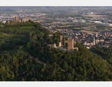 Veduta aerea di un castello medievale in pietra con mura e torri, situato su una collina boscosa. Sullo sfondo si estende una pianura con campi agricoli e un’area urbana con edifici e strade.
