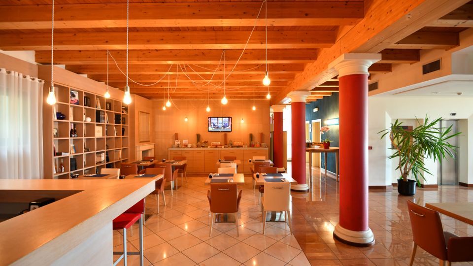 Interno di una sala ristorante o lounge con soffitto in travi di legno a vista, lampadine sospese e tavoli disposti su pavimento piastrellato lucido. A sinistra si vede un bancone e una libreria a parete, mentre a destra ci sono colonne rosse con capitelli bianchi, un corridoio e una pianta in vaso.