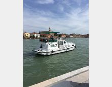 Imbarcazione bianca con tettuccio verde che naviga su un canale, vista dal bordo di una banchina in pietra. Sullo sfondo si vedono case basse colorate lungo la riva e un campanile di chiesa sotto un cielo parzialmente nuvoloso.
