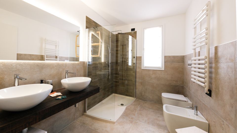 Bagno moderno con piastrelle beige e doccia angolare in vetro, doppio lavabo su piano scuro con grande specchio illuminato. Presenti finestra, termoarredo a parete, WC e bidet su pavimento in gres.
