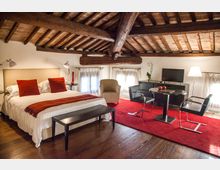 Interno di una camera in mansarda con soffitto in travi di legno e mattoni a vista, pavimento in legno e grandi finestre con tende bianche. Nella stanza ci sono un letto matrimoniale con cuscini rossi, una panca, poltrone e un’area con tavolo, sedie e mobile con TV su un tappeto rosso.