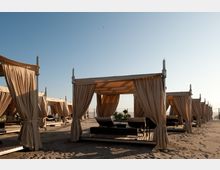 File di gazebo/cabane da spiaggia con tende beige su pedane di legno, dotati di lettini e tavolini, disposti sulla sabbia. Sullo sfondo si vede il mare e un cielo sereno.