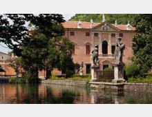 Villa storica con facciata rosa e tetto in coppi, affacciata su un canale con sponda in pietra. In primo piano si vedono statue su piedistalli lungo il bordo dell’acqua e alberi che incorniciano il giardino.