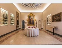 Sala museale in stile classico con pavimento in pietra e soffitto a cassettoni illuminato, con un grande ritratto in cornice dorata sulla parete di fondo. Al centro c’è un tavolo rotondo apparecchiato, mentre lungo le pareti laterali si vedono teche illuminate con porcellane e un grande specchio decorato.