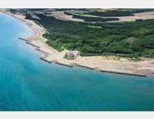 Vista aerea di una costa con ampia spiaggia sabbiosa e mare turchese, protetta da scogliere frangiflutti parallele alla riva. Dietro la spiaggia si estende una pineta fitta con un piccolo edificio vicino alla riva e campi agricoli nell’entroterra.