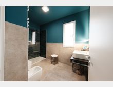Bagno moderno con pareti color petrolio e rivestimento in piastrelle beige, pavimento in gres e finestra che illumina l’ambiente. A sinistra c’è una doccia con parete in vetro e piastrelle scure, mentre a destra un lavabo rettangolare su mobile con ripiano e sgabelli in metallo.