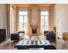 Ampio soggiorno luminoso con soffitto a travi in legno, due grandi finestre con tende beige e camino in marmo al centro della parete. Arredi moderni in pelle nera attorno a un tavolino in vetro su tappeto, con pavimento in terrazzo e vista su edifici storici attraverso le finestre.