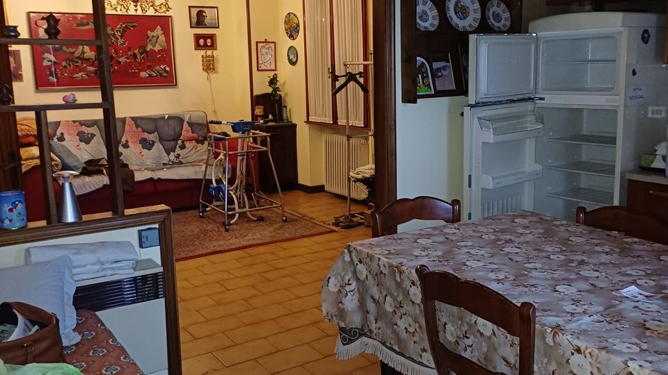 Interno di un appartamento con cucina-soggiorno open space, pavimento in piastrelle e tavolo da pranzo con tovaglia floreale accanto a un frigorifero aperto e mensole a parete. Un separé a scaffali divide la zona cucina dalla zona salotto, dove si vedono divano, quadri, lampadario e una porta-finestra.