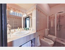 Bagno moderno con rivestimento in piastrelle chiare e decoro geometrico, lavabo incassato su ampio piano con grande specchio e luci. A destra si vedono bidet e WC e una doccia angolare con box in vetro.