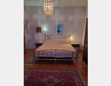 Camera da letto con letto matrimoniale in ottone al centro, comodini con lampade ai lati e lampadario a gocce sospeso. Pareti chiare con quadri, pavimento in parquet e tappeti decorati in primo piano.