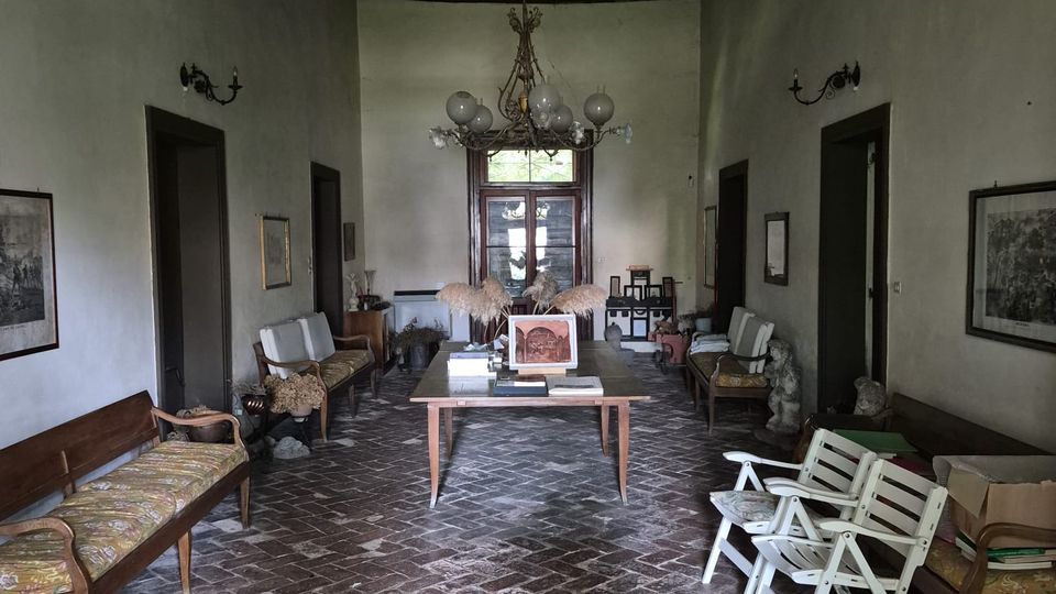 Ampio salone interno con pavimento in mattoni a spina di pesce, pareti intonacate chiare e soffitto alto con travi in legno a vista. Al centro ci sono un tavolo e un lampadario, con panche e sedie lungo i lati e una porta vetrata in fondo alla stanza.
