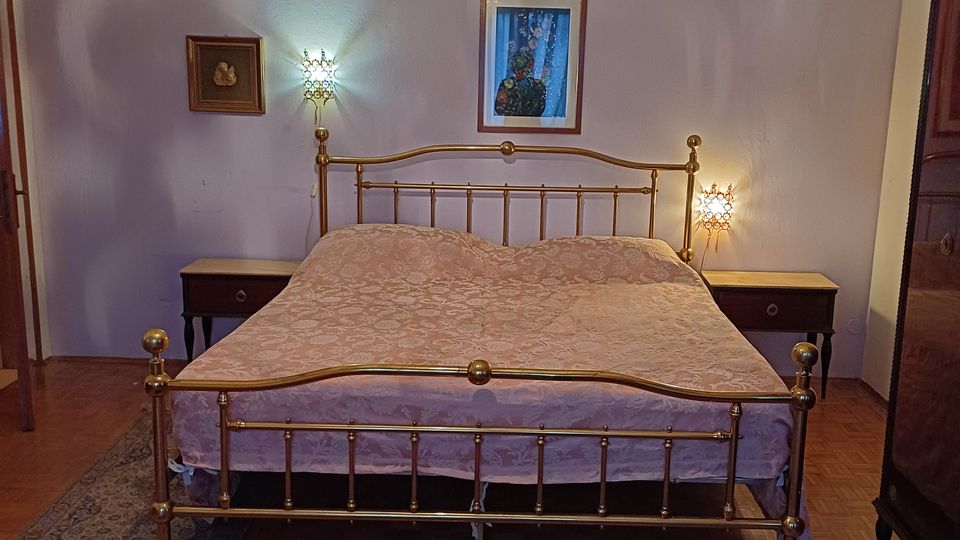 Camera da letto con letto matrimoniale in ottone al centro, comodini con lampade ai lati e lampadario a gocce sospeso. Pareti chiare con quadri, pavimento in parquet e tappeti decorati in primo piano.