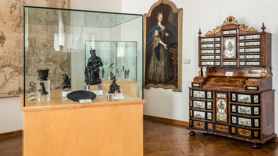 Sala museale con pavimento in parquet e pareti bianche, con una grande vetrina espositiva in vetro su base in legno contenente piccole sculture. Sulla parete sono presenti una grande mappa antica e un ritratto incorniciato, accanto a un elaborato mobile-scrivania intarsiato con numerosi cassetti decorati.