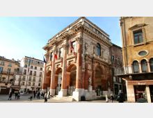 Edificio storico in piazza con facciata in mattoni e pietra, portico ad arcate e colonne, balconata decorata e statue nelle nicchie. Intorno si vedono palazzi multipiano con finestre e balconi che delimitano lo spazio urbano.
