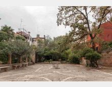 Cortile lastricato in pietra con aiuole e alberi maturi, con una piccola vasca o pozzo in pietra al centro. Intorno si vedono panchine lungo il muro, vegetazione fitta e edifici con tetti e antenne sullo sfondo, oltre a una parete in mattoni rossi su un lato.