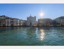 Veduta di un canale urbano con una chiesa in pietra chiara al centro e palazzi storici a più piani lungo le rive. In primo piano l’acqua del canale con riflessi del sole e un pontile per vaporetti davanti alla chiesa.