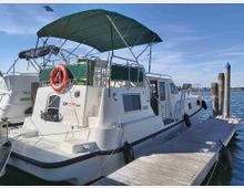 Barca a motore bianca ormeggiata a un pontile di legno in un porto, con tendalino verde e salvagente arancione a poppa. Sullo sfondo si vedono pali d’ormeggio e acqua calma sotto un cielo sereno.