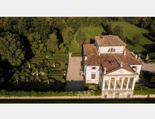 Veduta aerea di una villa storica con facciata a portico colonnato e tetti in coppi, affiancata da un giardino formale geometrico con siepi potate, statue e fontana centrale. Intorno si estendono viali ghiaiosi, prati e alberi ad alto fusto.