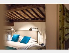 Camera da letto in stile rustico con soffitto a travi di legno a vista, letto matrimoniale con testiera imbottita e cuscini blu. Ai lati del letto ci sono lampade da lettura e piccoli comodini, con tende chiare alle finestre.