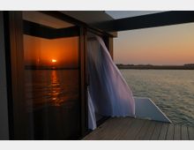 Terrazza in legno di una struttura moderna sul bordo dell’acqua, con pareti scure e una grande porta vetrata. Una tenda bianca svolazza verso l’esterno e il tramonto si riflette sul vetro e sulla superficie del mare.