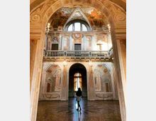 Interno di un palazzo storico con grande arco in pietra e sala decorata da affreschi, colonne e balconata superiore con finestre ad arco. Sul fondo si vede un alto portale illuminato e un pavimento lucido che riflette la luce.