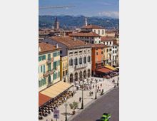 Veduta dall’alto di una grande piazza urbana con caffè all’aperto sotto tende color ocra, lampioni e alberi in vaso, circondata da edifici storici con facciate color pastello e tetti in terracotta. Sullo sfondo si vedono campanili e colline verdi sotto un cielo sereno.