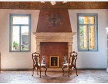 Salone d’epoca con grande camino in pietra e cappa decorata al centro, affiancato da due finestre con infissi azzurri. Pavimento in cotto e due sedie in legno intagliato davanti al focolare, con vasi e un quadro appoggiati alla base.