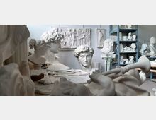 Interno di un laboratorio di scultura pieno di calchi in gesso, con busti classici su un tavolo e su scaffalature. Sullo sfondo si vedono rilievi e statue appoggiate alle pareti in un ambiente luminoso e affollato di opere.