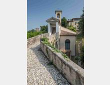 Chiesetta su una collina con campanile e portico neoclassico, raggiunta da una rampa in ciottoli con muretto in pietra. Intorno si vedono alberi e edifici storici sullo sfondo sotto un cielo sereno.