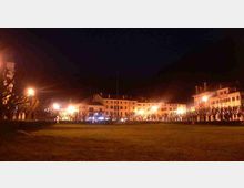 Veduta notturna di una piazza con grande prato in primo piano e filari di alberi potati lungo i margini, illuminati da lampioni. Sullo sfondo si vedono edifici storici a più piani con facciate chiare e finestre regolari, illuminati dalle luci stradali.