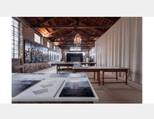 Interno di un grande loft industriale con pareti in mattoni a vista e ampie finestre a griglia, soffitto alto in legno con travi e lampadari sospesi. Tavoli da lavoro con stampe e disegni, pannelli artistici lungo la parete sinistra e una lunga tenda chiara che divide lo spazio sul lato destro.