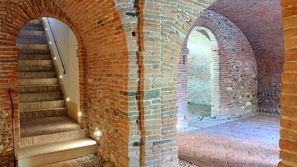 Interno di un ambiente storico con volte e archi in mattoni a vista, pavimento in cotto e dettagli in ciottoli. Una scala illuminata conduce a un livello superiore.