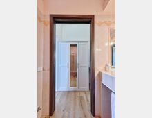 Vista da un bagno piastrellato con cornice in legno scuro verso un corridoio con pavimento in legno e armadi bianchi a tutta parete. A destra si vede il lavabo con specchio e piano d’appoggio, mentre in fondo si intravede una zona doccia.