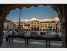Veduta di una piazza storica attraverso un grande arco in pietra, con pavimentazione in ciottoli e una fila di massi su una struttura metallica in primo piano. Sullo sfondo si vedono edifici porticati e un palazzo civico con orologio centrale e balconi, sotto un cielo con nuvole.