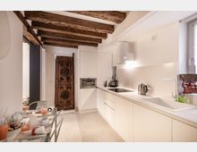 Cucina moderna e compatta con mobili bianchi a linea continua, piano cottura a induzione, lavello e cappa cilindrica, illuminata da una finestra laterale. Soffitto con travi in legno a vista e piccolo tavolo da pranzo in vetro nell’angolo, con un pannello decorativo in legno appoggiato alla parete.