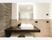 Bagno moderno con lavabo rettangolare in ceramica su mensola in legno scuro, grande specchio e pareti rivestite in piastrelle beige e effetto legno. Sullo sfondo si vede un termoarredo bianco e una cabina doccia in vetro.