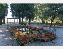 Giardino alberato con vialetti in ghiaia e una vasca rettangolare con ninfee e piccola fontana centrale, circondata da aiuole di fiori rossi e recinzione in ferro. Sullo sfondo si vede una terrazza con balaustra in pietra e una struttura a colonne affacciata sul paesaggio.