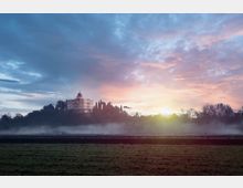 Edificio storico con cupola su una collina, circondato da alberi, visto da un campo in primo piano con una fascia di nebbia bassa. Sullo sfondo cielo all’alba con nuvole e luce dorata vicino all’orizzonte.